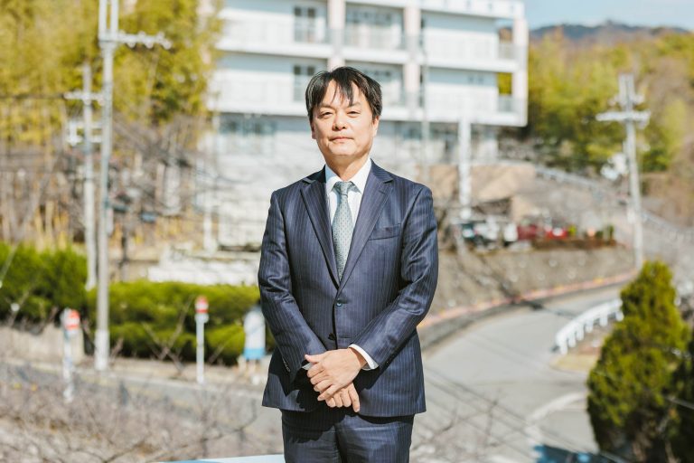 施設前の庭園で、紺のスーツ姿の男性スタッフが立っているポートレート写真